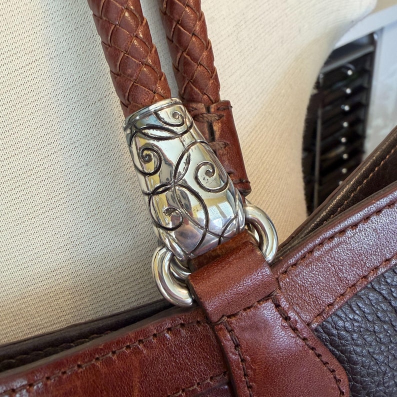 May include: Close-up of a brown leather handbag with a braided handle. The handle is attached to the bag with a silver-colored metal clasp with a decorative swirl design. The bag has a textured leather body.