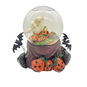 May include: A Halloween snow globe with two ghosts, pumpkins, and a tombstone that reads "RIP". The snow globe is sitting on a purple fabric base with more pumpkins.
