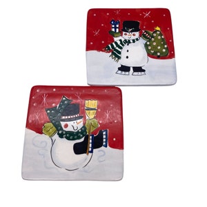 May include: Two red and white ceramic plates with snowman designs. One snowman is holding a Christmas tree and the other is holding a broom.