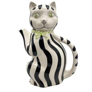 May include: A ceramic teapot shaped like a cat, featuring a white base with bold black stripes. The cat's face is white with green eyes and black markings. A green and white bow adorns the neck. The teapot has a handle and spout.