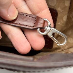 May include: Close-up of a brown leather strap with a silver-colored metal clasp. The strap is stitched and appears to be part of a bag or purse. The clasp is open, revealing the interior of the bag.