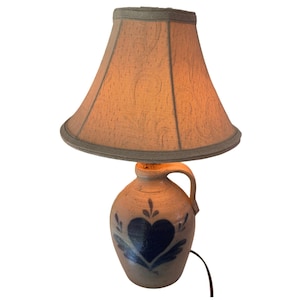 May include: A table lamp featuring a beige ceramic jug base with a dark blue painted heart and leaf motif. The lamp has a single side handle and is topped with a flared, textured beige fabric lampshade that emits a warm, soft glow.