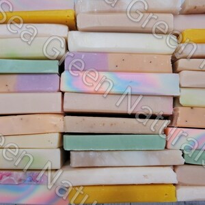 May include: A collection of handmade soap bars in various colors and patterns. The soaps are stacked together, showcasing a variety of hues including yellow, green, purple, and pink. Some bars have speckled textures, while others display marbled designs.