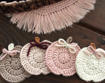 May include: Four handmade crochet pumpkin coasters in shades of beige, pink, and white, each with a stem and bow detail. The coasters are arranged on a wooden surface, with a woven basket and fringed fabric in the background.
