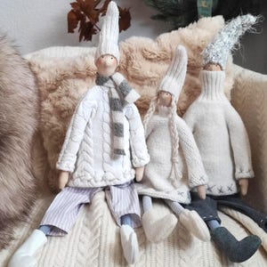 Puede incluir: Tres muñecas hechas a mano vestidas con ropa de invierno. Las muñecas visten suéteres y gorros de punto blancos, con bufandas y pantalones a rayas. Una muñeca tiene una bufanda a rayas grises y blancas. Las muñecas están sentadas sobre una superficie texturizada de color crema.