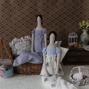 May include: Two handmade fabric dolls dressed in vintage-inspired clothing. The doll on the left is wearing a blue and white striped dress. The doll on the right is wearing a white dress with blue floral print.