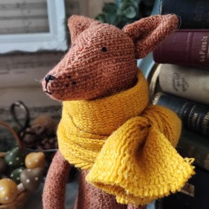 May include: A brown knitted fox toy wearing a yellow knitted scarf. The fox is sitting on a shelf with books behind it.