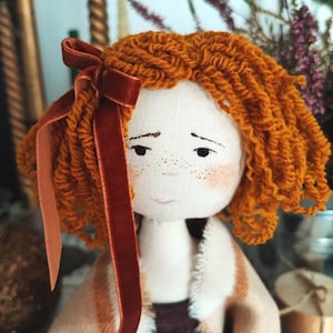 May include: A handmade fabric doll with orange yarn hair and a dark red velvet bow. The doll has a painted face with freckles and is wearing a tan and brown coat. The doll's clothing is a mix of tan, brown, and burgundy fabrics.