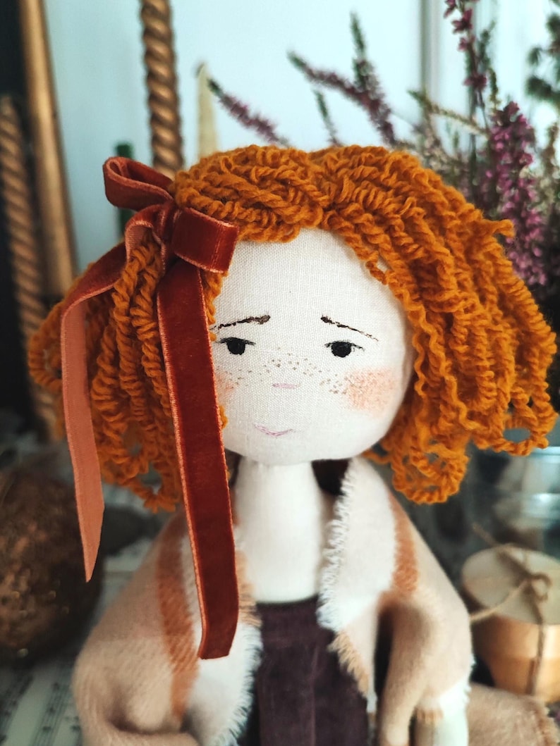 May include: A handmade fabric doll with orange yarn hair and a dark red velvet bow. The doll has a painted face with freckles and is wearing a tan and brown coat. The doll's clothing is a mix of tan, brown, and burgundy fabrics.