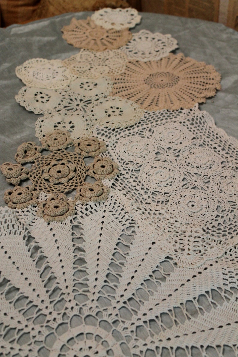 May include: A collection of white and cream crocheted doilies of various sizes and patterns arranged on a gray surface.