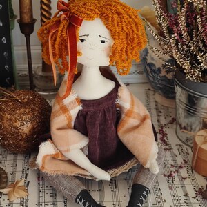 May include: A handmade doll with orange yarn hair and a velvet bow. The doll wears a burgundy dress, a plaid shawl, and gray leggings. Black boots with white lacing complete the outfit. The doll is sitting on a surface with sheet music.