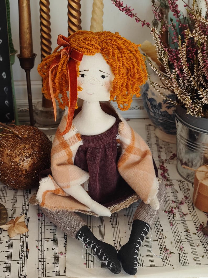 May include: A handmade doll with orange yarn hair and a velvet bow. The doll wears a burgundy dress, a plaid shawl, and gray leggings. Black boots with white lacing complete the outfit. The doll is sitting on a surface with sheet music.