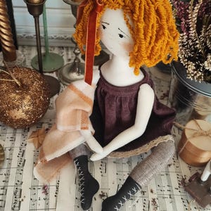 May include: A handmade doll with orange yarn hair and a burgundy dress. The doll has a white face with freckles and is wearing black boots with white stitching. A peach and cream colored scarf is draped over the doll's arm.