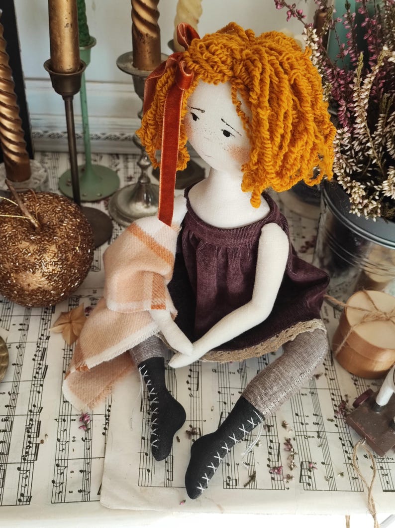 May include: A handmade doll with orange yarn hair and a burgundy dress. The doll has a white face with freckles and is wearing black boots with white stitching. A peach and cream colored scarf is draped over the doll's arm.