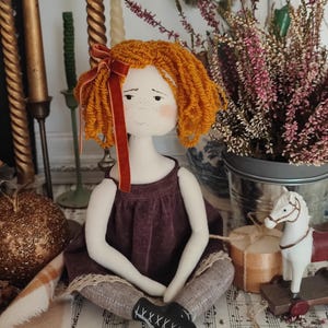 May include: A handmade doll with orange yarn hair and a burgundy dress. The doll has a white face with freckles and is wearing gray leggings and black shoes. A red velvet ribbon adorns the doll's hair. The doll is sitting on sheet music.