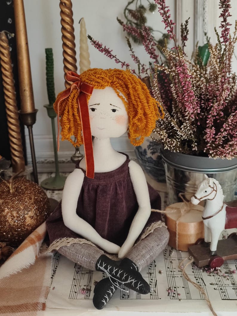 May include: A handmade doll with orange yarn hair and a burgundy dress. The doll has a white face with freckles and is wearing gray leggings and black shoes. A red velvet ribbon adorns the doll's hair. The doll is sitting on sheet music.