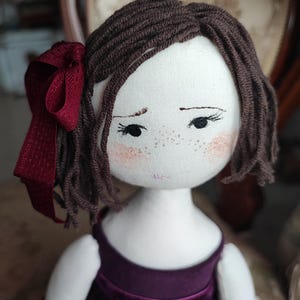 May include: A handmade cloth doll with brown yarn hair and a burgundy ribbon. The doll has a white face with embroidered features, including freckles and rosy cheeks. It is wearing a dark purple velvet dress.