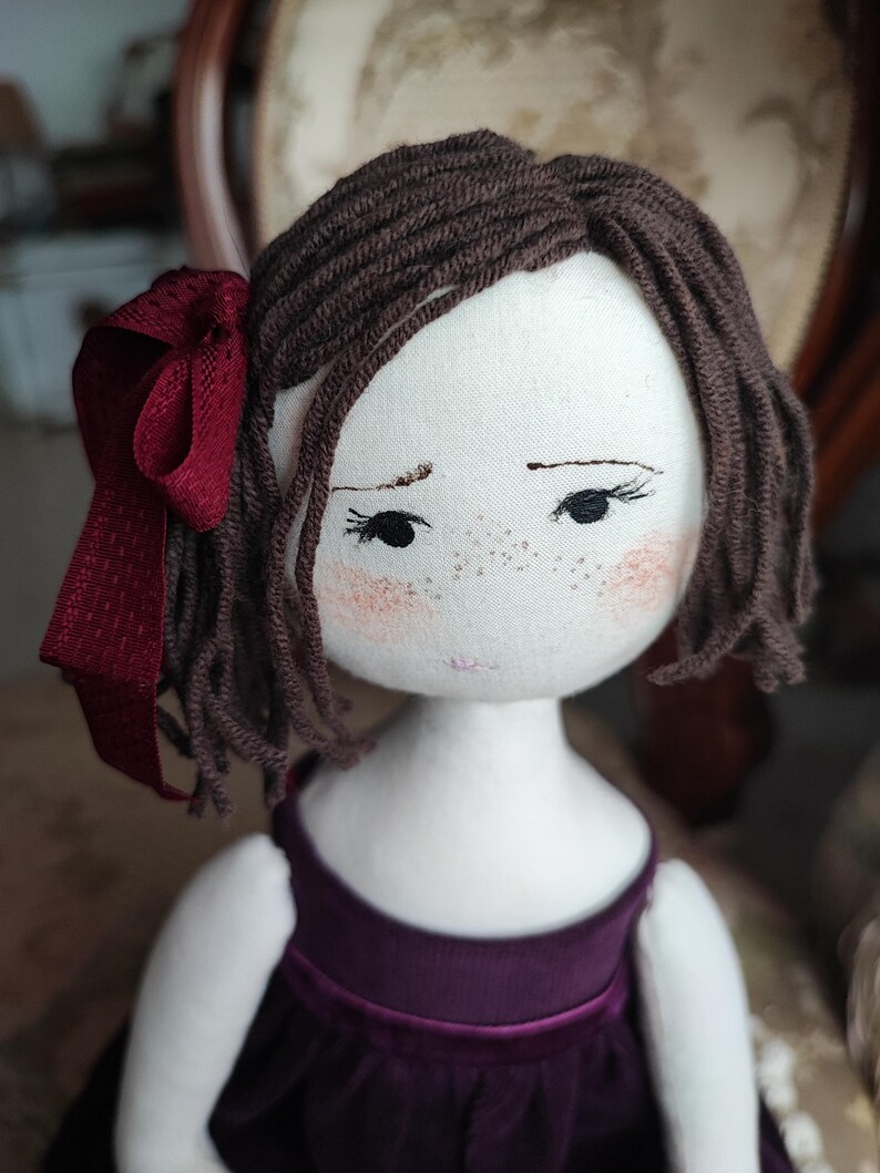May include: A handmade cloth doll with brown yarn hair and a burgundy ribbon. The doll has a white face with embroidered features, including freckles and rosy cheeks. It is wearing a dark purple velvet dress.