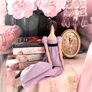 May include: A purple and white striped dress doll sitting on a pink floral tablecloth with a stack of books and a gold clock with Roman numerals.