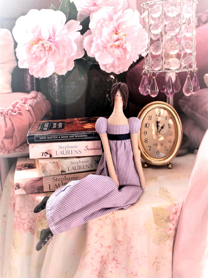 May include: A purple and white striped dress doll sitting on a pink floral tablecloth with a stack of books and a gold clock with Roman numerals.