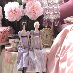 May include: Two dolls dressed in blue and white striped dresses sit on a table with a pink tablecloth. The dolls have brown and blonde hair and are wearing white lace trim on their dresses. A pink floral arrangement is in the background.