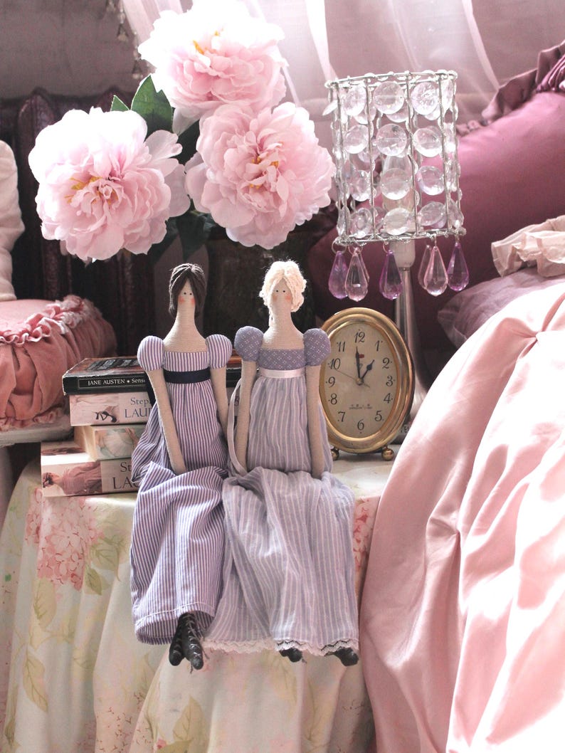 May include: Two dolls dressed in blue and white striped dresses sit on a table with a pink tablecloth. The dolls have brown and blonde hair and are wearing white lace trim on their dresses. A pink floral arrangement is in the background.