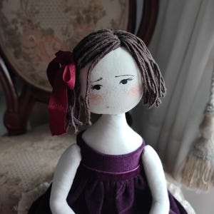 May include: Handmade cloth doll with brown yarn hair and a burgundy ribbon. The doll has a white face with painted features, including freckles and rosy cheeks. It wears a dark purple velvet dress.