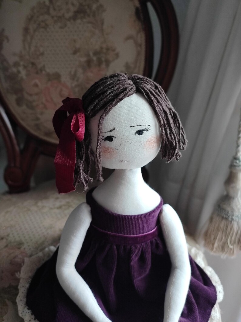 May include: Handmade cloth doll with brown yarn hair and a burgundy ribbon. The doll has a white face with painted features, including freckles and rosy cheeks. It wears a dark purple velvet dress.
