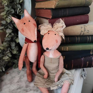 May include: Two handmade plush fox dolls, one standing and one sitting. The standing fox is wearing a red scarf and has a brown nose. The sitting fox is wearing a beige dress with lace trim and a white lace bow. Both foxes have pink faces and black eyes.
