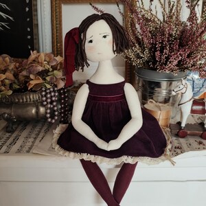 May include: A handmade cloth doll with a burgundy velvet dress and matching tights. The doll has brown hair, a red bow, and silver-tipped shoes. The doll is sitting on a white surface, with a vintage aesthetic.