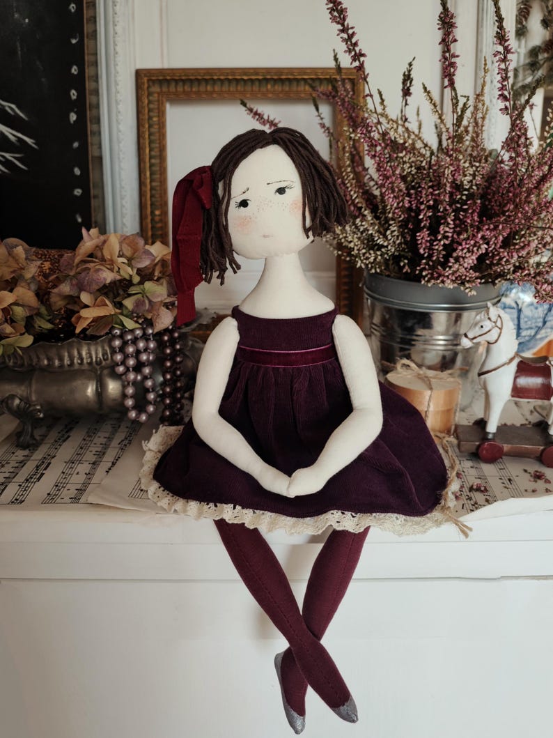 May include: A handmade cloth doll with a burgundy velvet dress and matching tights. The doll has brown hair, a red bow, and silver-tipped shoes. The doll is sitting on a white surface, with a vintage aesthetic.