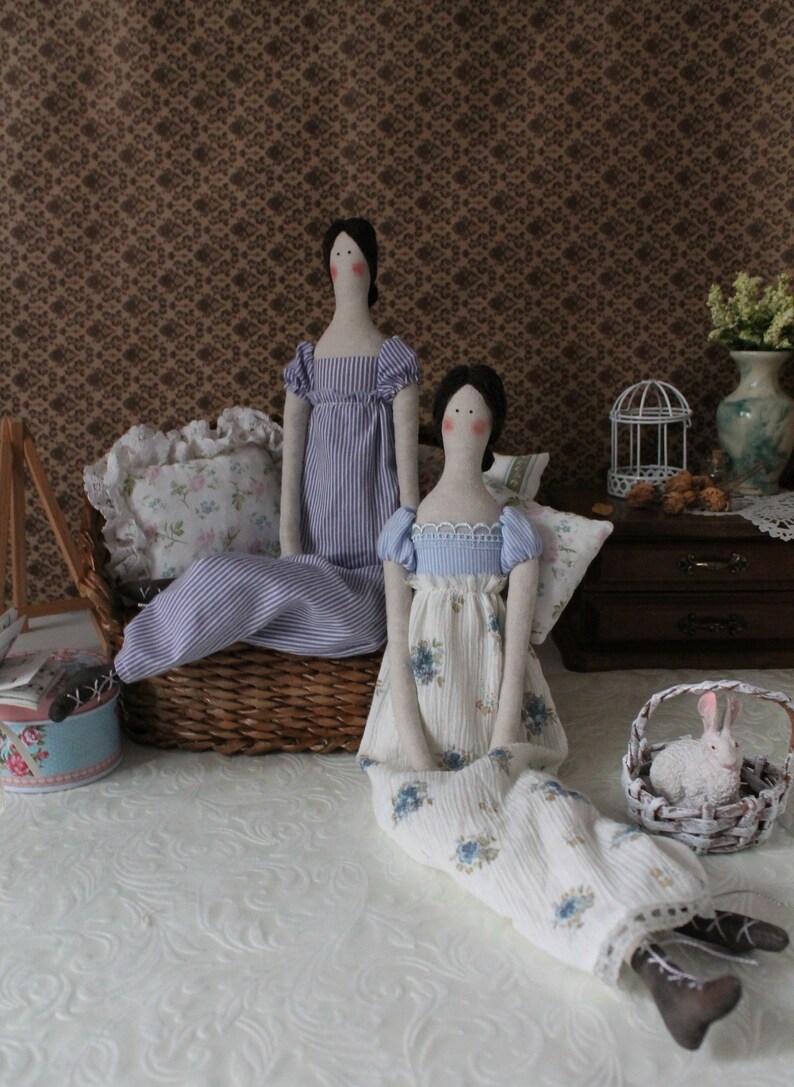 May include: Two handmade fabric dolls dressed in vintage-inspired clothing. The doll on the left is wearing a blue and white striped dress. The doll on the right is wearing a white dress with blue floral print.
