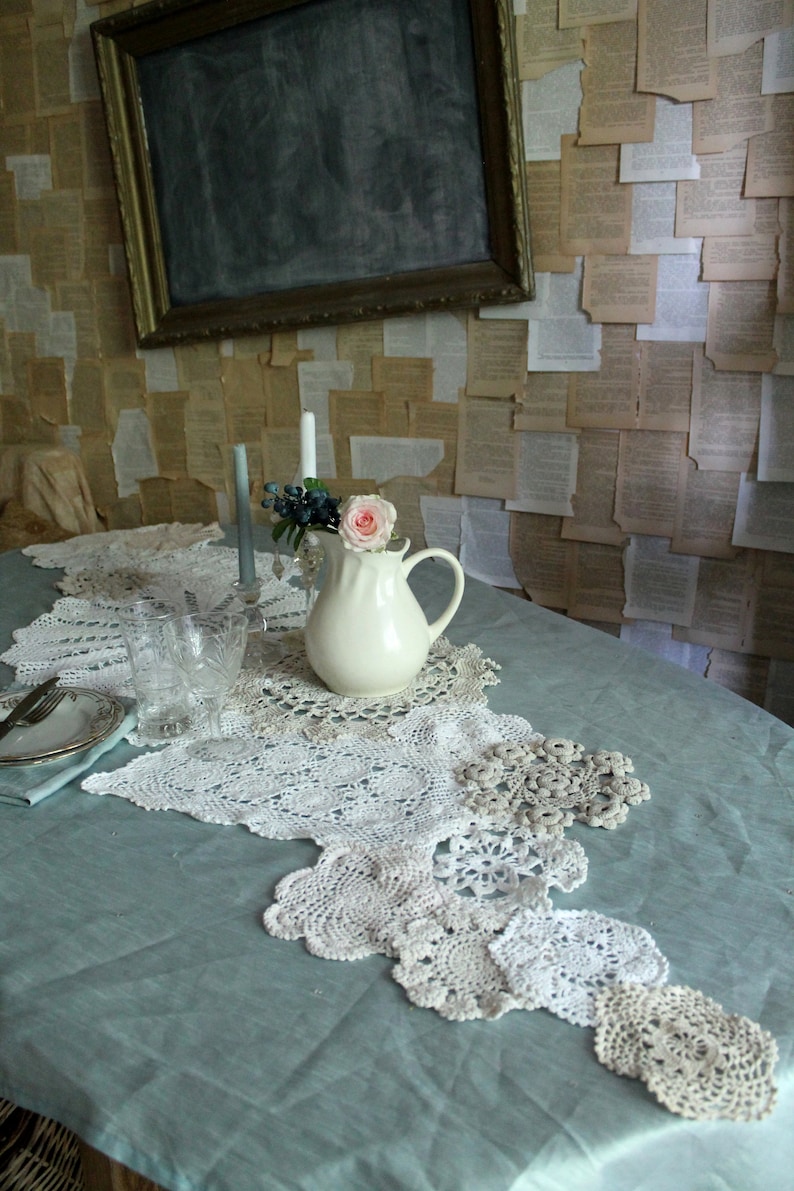 May include: A white ceramic pitcher with a pink flower sits on a table with a blue tablecloth and a white crocheted table runner. The table runner is made of several different doilies.