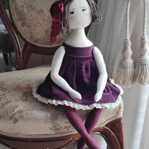 May include: A handmade cloth doll with a white face, short brown hair, and a burgundy bow. The doll wears a purple dress with white lace trim and matching leggings. It is seated on a vintage floral patterned chair.