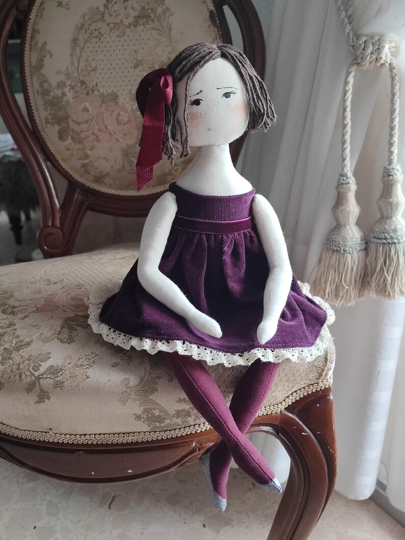 May include: A handmade cloth doll with a white face, short brown hair, and a burgundy bow. The doll wears a purple dress with white lace trim and matching leggings. It is seated on a vintage floral patterned chair.