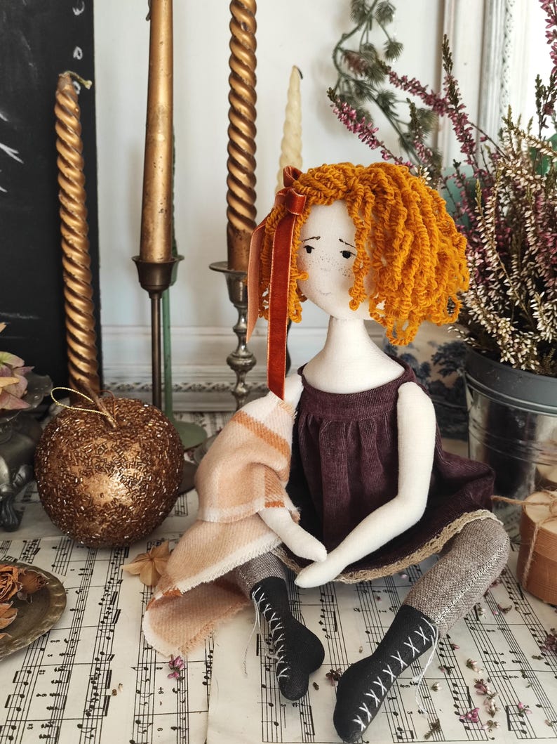 May include: A handmade doll with orange yarn hair, wearing a burgundy dress and black boots with white lacing. The doll is seated on sheet music, with a small blanket and a decorative gold ornament nearby. Several gold candles and a floral arrangement are in the background.