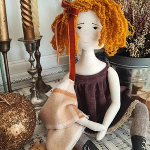 May include: A handmade doll with orange yarn hair and a burgundy dress. The doll has a cream-colored face with freckles and is holding a tan and cream-colored scarf. The doll is wearing black boots with white laces.