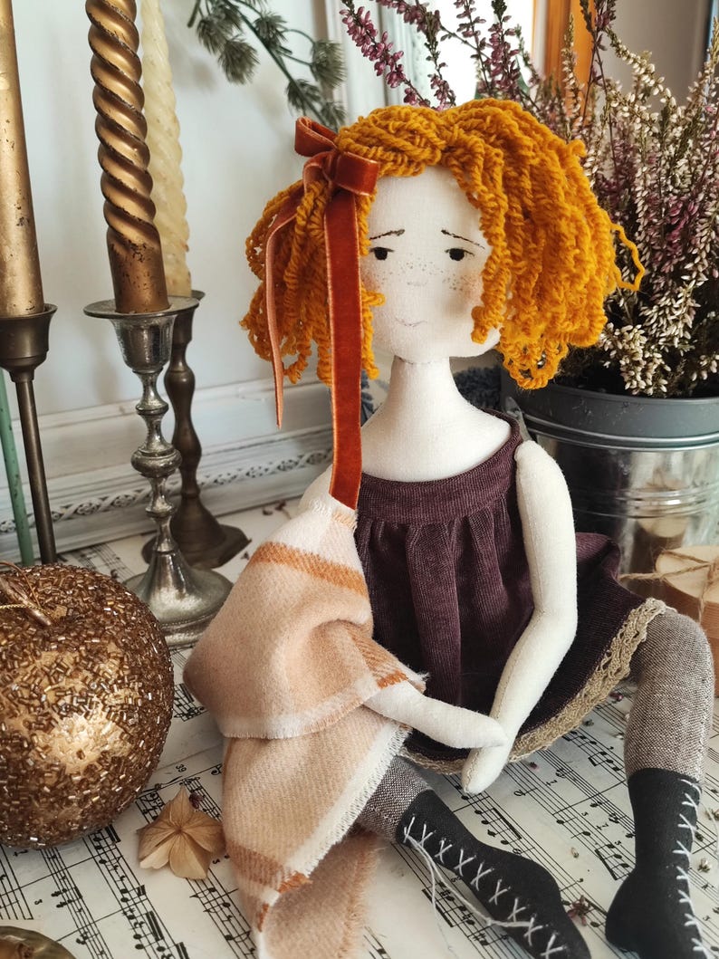 May include: A handmade doll with orange yarn hair and a burgundy dress. The doll has a cream-colored face with freckles and is holding a tan and cream-colored scarf. The doll is wearing black boots with white laces.