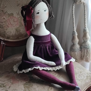 May include: A handmade cloth doll with a serene expression. The doll wears a deep purple velvet dress with a white lace trim, matching tights, and a burgundy bow in her short brown hair. The doll is seated on a floral patterned chair.