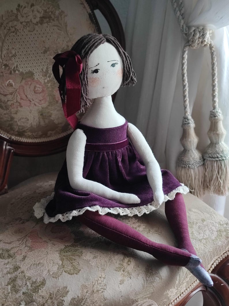 May include: A handmade cloth doll with a serene expression. The doll wears a deep purple velvet dress with a white lace trim, matching tights, and a burgundy bow in her short brown hair. The doll is seated on a floral patterned chair.