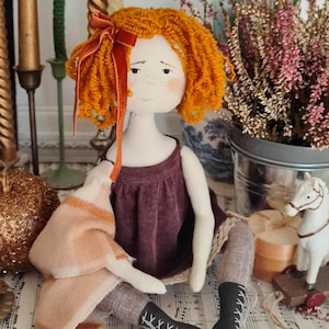 May include: A handmade doll with orange yarn hair and a burgundy dress. The doll is wearing gray stockings and black boots with white laces. A peach and white plaid blanket is draped over the doll's arm. A red velvet bow adorns the doll's hair.