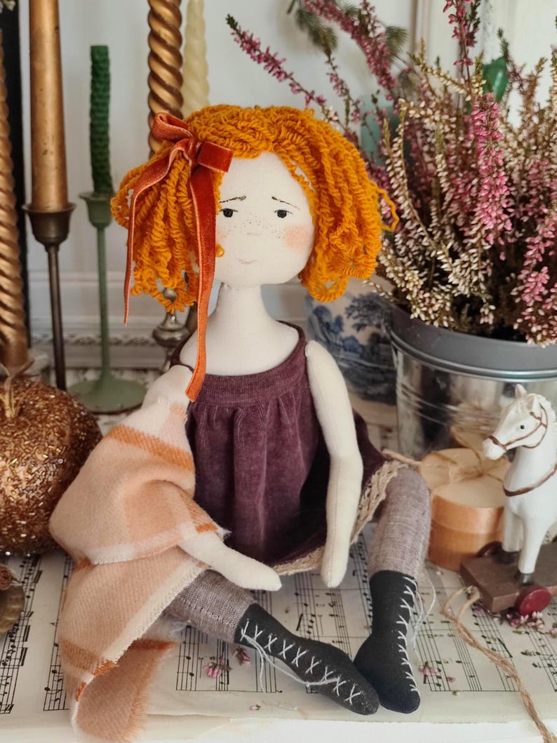 May include: A handmade doll with orange yarn hair and a burgundy dress. The doll is wearing gray stockings and black boots with white laces. A peach and white plaid blanket is draped over the doll's arm. A red velvet bow adorns the doll's hair.