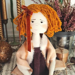 May include: A handmade fabric doll with orange yarn hair and a velvet bow. The doll wears a burgundy dress and a shawl with peach and cream stripes. The doll has freckles and is set against a backdrop of candles and flowers.