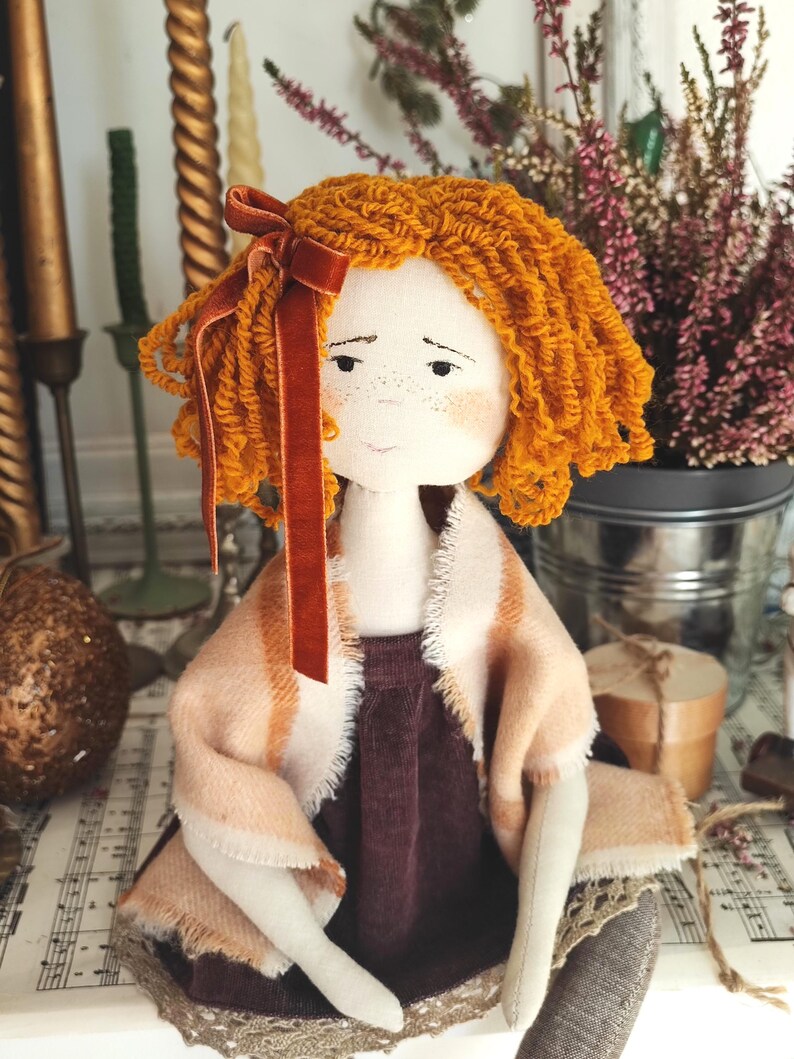 May include: A handmade fabric doll with orange yarn hair and a velvet bow. The doll wears a burgundy dress and a shawl with peach and cream stripes. The doll has freckles and is set against a backdrop of candles and flowers.