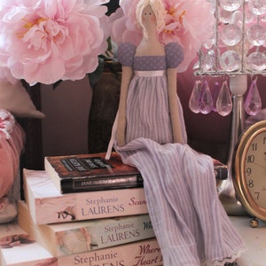 May include: A stack of books with a doll sitting on top. The doll is wearing a lavender dress with white stripes and a white lace trim. The doll has blonde hair and a pink flower behind it. The books are all by the same author, Stephanie Laurens. The titles of the books are "Scan", "Where Heart Leads", and "The Wanderer".