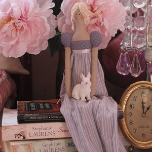 May include: A porcelain doll wearing a lavender dress with white stripes sits on a stack of books. The doll has blonde hair and is holding a white rabbit figurine. The books have titles by Stephanie Laurens.