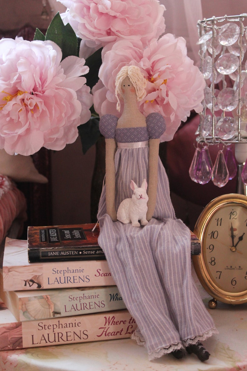 May include: A porcelain doll wearing a lavender dress with white stripes sits on a stack of books. The doll has blonde hair and is holding a white rabbit figurine. The books have titles by Stephanie Laurens.