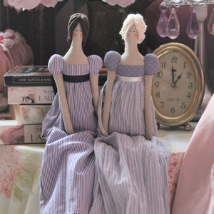 May include: Two fabric dolls dressed in purple and white striped dresses with white lace trim. The dolls are sitting on a table with a pink tablecloth and a stack of books. A gold clock is on the table behind the dolls.