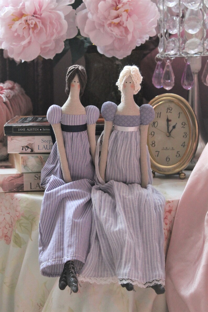 May include: Two fabric dolls dressed in purple and white striped dresses with white lace trim. The dolls are sitting on a table with a pink tablecloth and a stack of books. A gold clock is on the table behind the dolls.