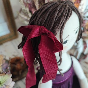 May include: A handmade doll with brown yarn hair and a burgundy ribbon tied in a bow. The doll is wearing a purple dress. The doll's face is painted with delicate features, including small eyes and freckles.